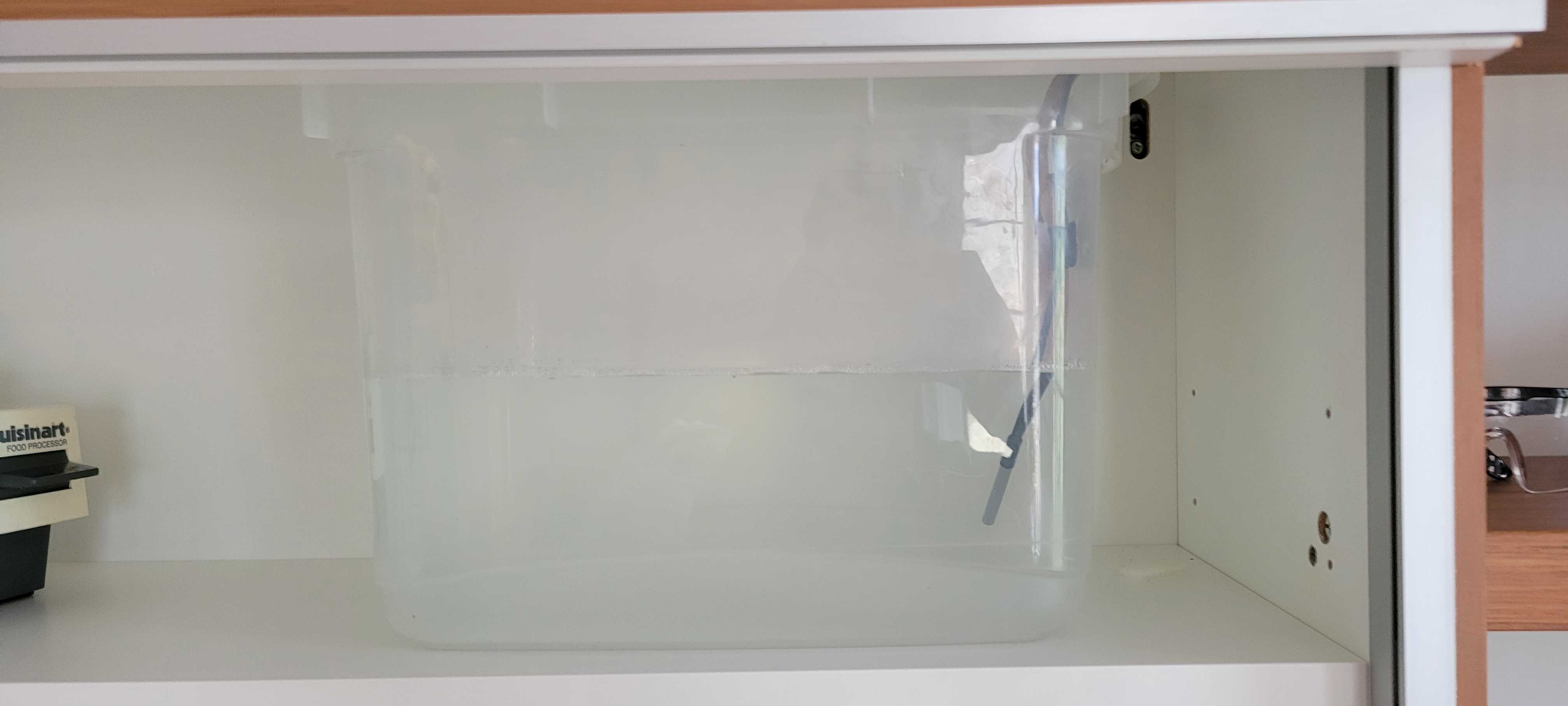 an uncovered plastic bucket filled with water sitting inside a cabinet. there are two latex tubes running from a hole in the top of the cabinet into the bucket, as well as a temperature probe